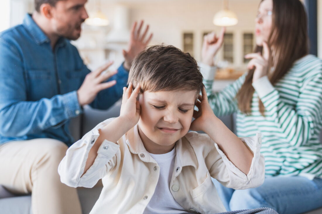 Young boy needing family law solicitor holds ears with face in grimace while parents argue in background